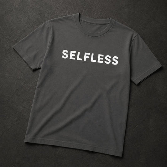 A dark gray crewneck t-shirt with the word 'SELFLESS' printed in white capital letters across the chest, laid flat on a dark textured background.