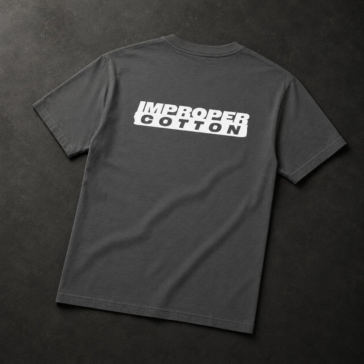 A dark gray crewneck t-shirt laid flat on a dark textured background, featuring a white graphic print on the back that reads 'IMPROPER COTTON' inside a tilted white rectangle.
