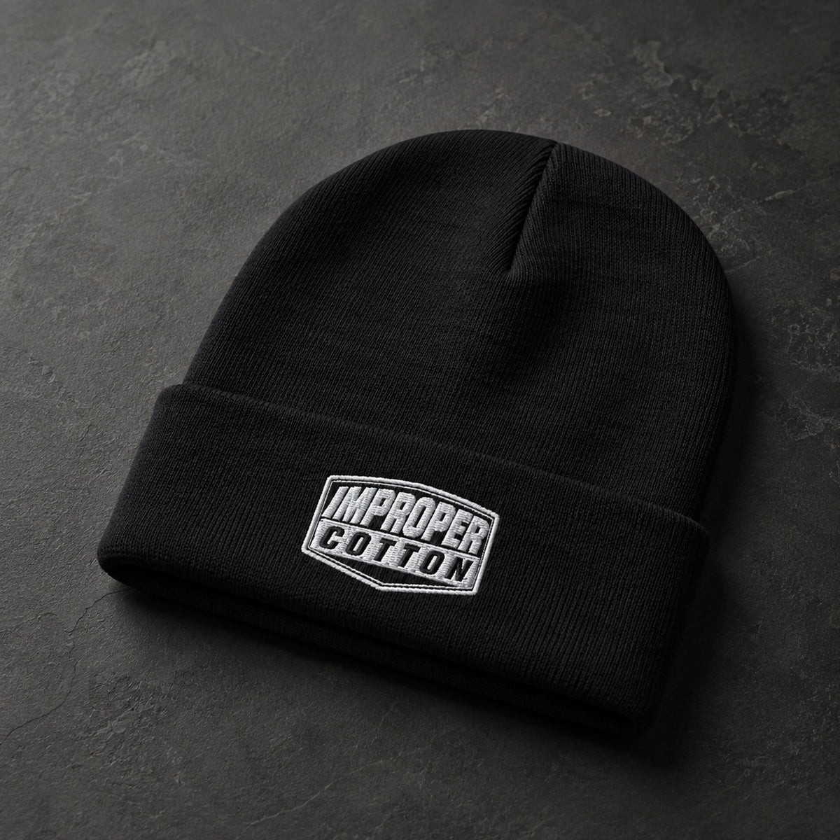 A black knit beanie with a folded cuff and an embroidered white and black 'IMPROPER COTTON' logo patch, displayed on a dark textured surface.