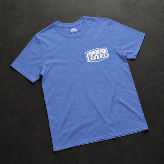 A heather blue crewneck t-shirt with a white graphic logo on the left chest reading 'IMPROPER COTTON', laid flat on a dark textured background.