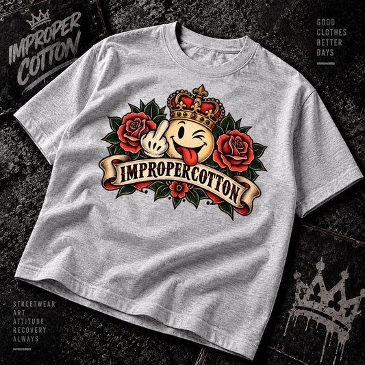A light heather gray graphic t-shirt laid flat on a dark textured background. The shirt features a large retro tattoo-style graphic in the center with red roses, a yellow face with a crown, and a banner reading 'IMPROPERCOTTON'.