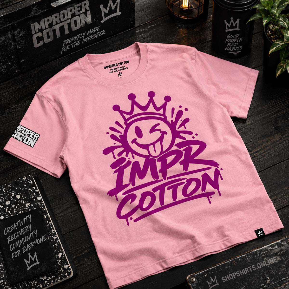A pink short-sleeve t-shirt with a large purple graphic on the front reading 'IMPR COTTON' beneath a smiling crown face. The shirt is laid flat on a dark wooden surface with branded black boxes, notebooks, and a candle around it.