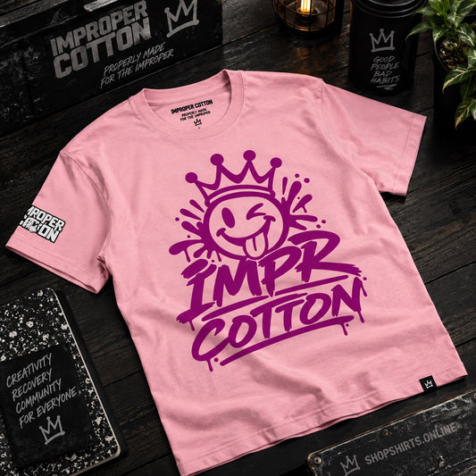 A pink short-sleeve t-shirt with a large purple graphic on the front reading 'IMPR COTTON' beneath a smiling crown face. The shirt is laid flat on a dark wooden surface with branded black boxes, notebooks, and a candle around it.