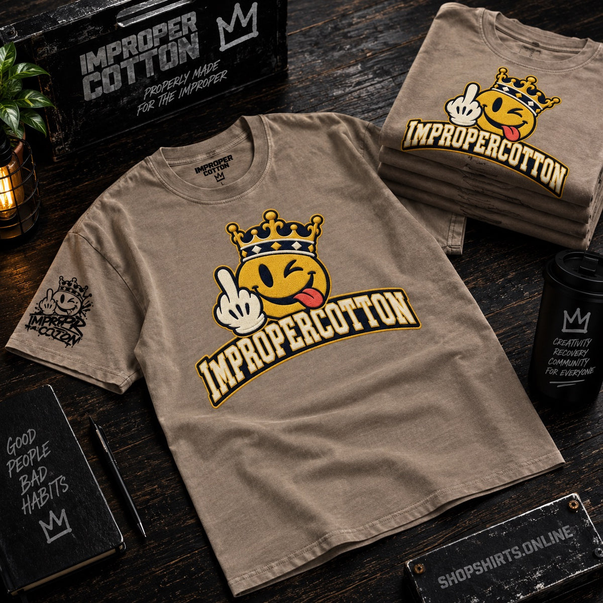A tan graphic T-shirt laid flat on a dark wooden surface, with a matching folded T-shirt in the background. The shirt features a winking emoji wearing a crown and the text 'IMPROPERCOTTON' in yellow outlined lettering.