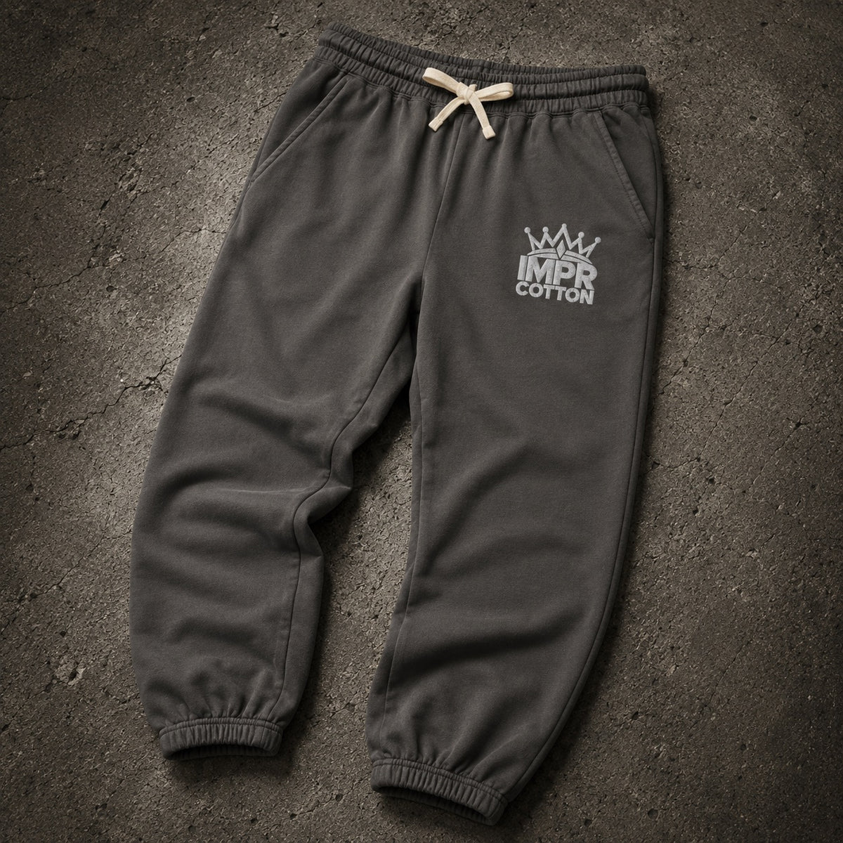 Dark charcoal gray sweatpants with a white 'IMPR COTTON' crown logo on the left thigh, featuring an elastic waistband with a cream drawstring and elastic cuffs, laid flat on a textured dark gray surface.