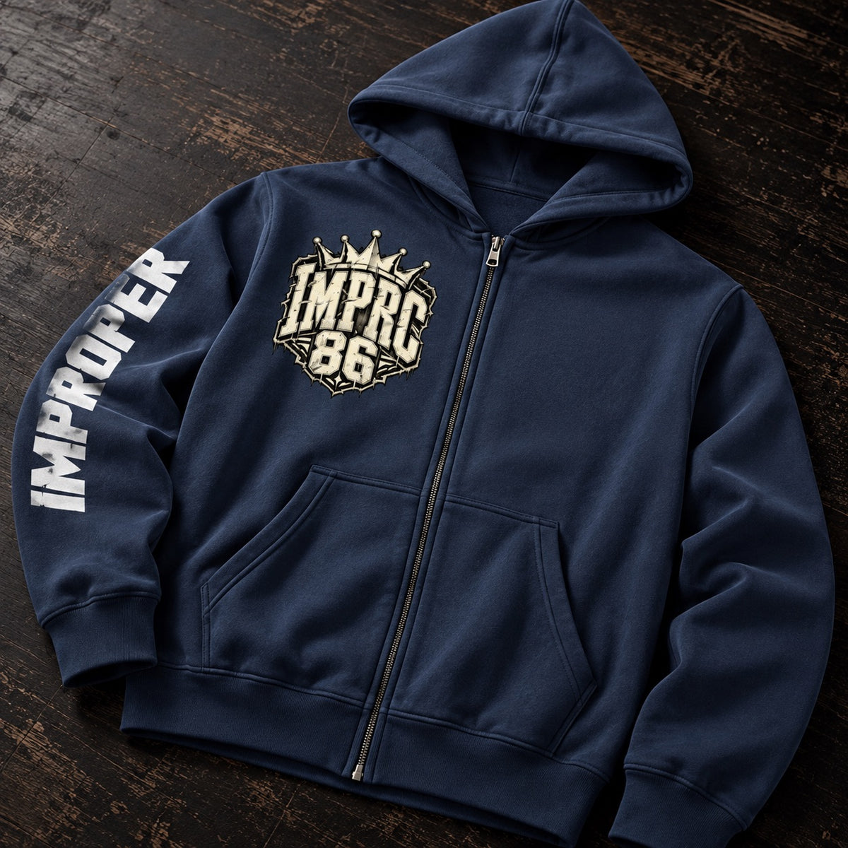 A navy blue zip-up hoodie laid flat on a dark wooden surface. The hoodie features a large distressed gold and white 'IMPRC 86' crest on the left chest with a small crown graphic, and the word 'IMPROPER' printed vertically in white on the right sleeve.