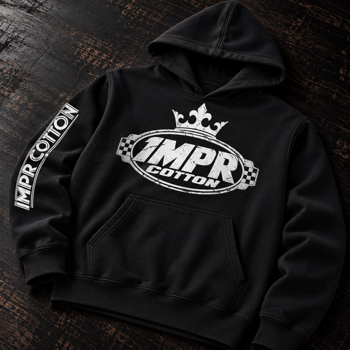 A black pullover hoodie laid flat on a dark wooden surface, featuring a large white '1MPR COTTON' graphic with a crown on the chest and '1MPR COTTON' printed down one sleeve.