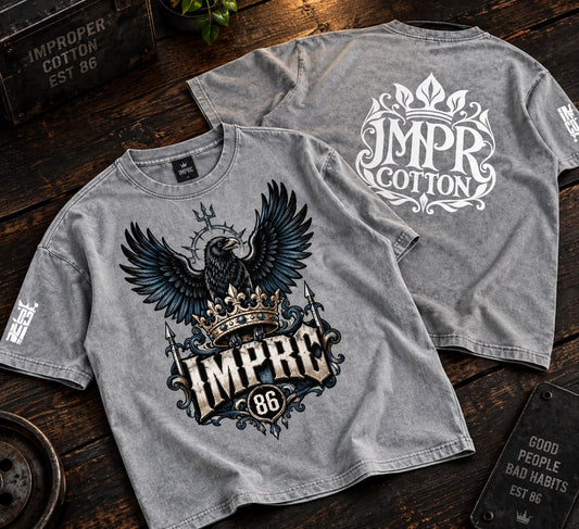 Two heather gray graphic T-shirts displayed on a dark wooden surface. The front shirt features a large black and white eagle with a gold crown and the text 'IMPRC 86'.