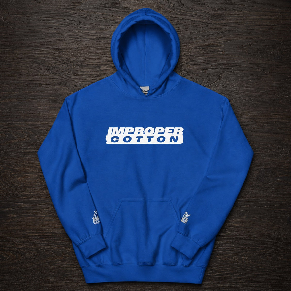 A royal blue pullover hoodie with a white graphic text print reading 'IMPROPER COTTON' across the chest, laid flat on a dark wood background.