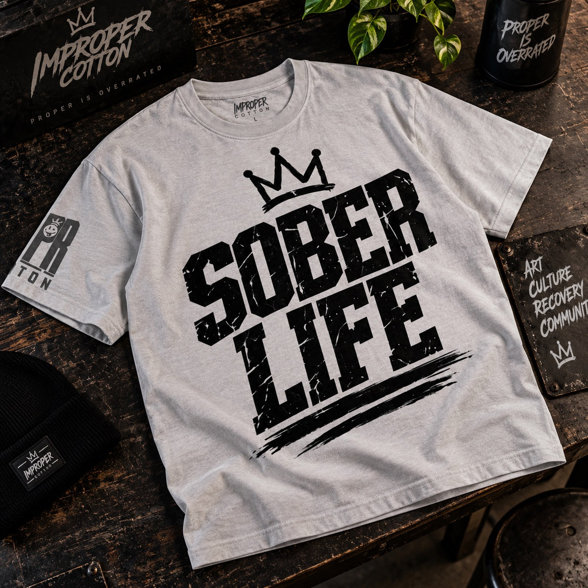 Light gray Improper Cotton Sober Life graphic T-shirt featuring distressed black lettering and crown design, laid slightly angled on a dark wooden table with branded streetwear accessories.