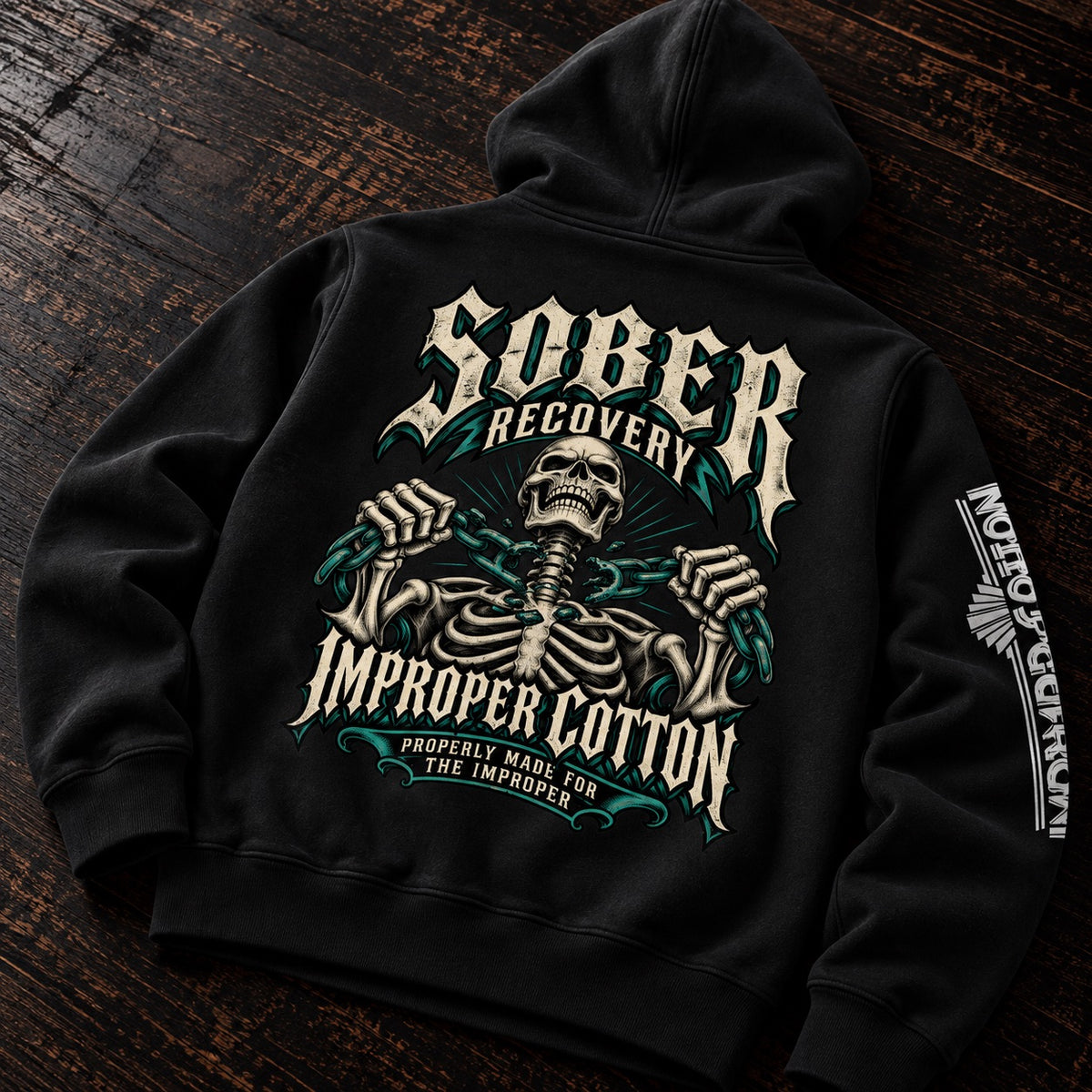 A black pullover hoodie laid flat on a dark wooden surface, featuring a large back graphic of a skeleton with the words 'Sober Recovery Improper Cotton' in distressed white and teal lettering. A smaller white graphic appears on the sleeve.