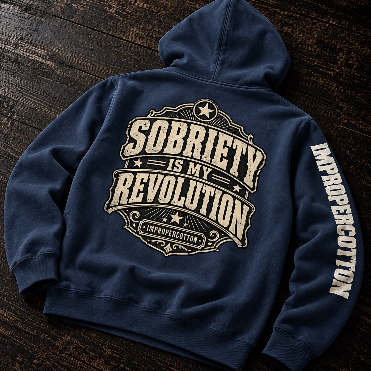 A navy blue hoodie laid flat on a dark wooden surface, showing the back design with a large cream graphic reading 'Sobriety is my Revolution' and 'Impropercotton,' plus matching text down one sleeve.
