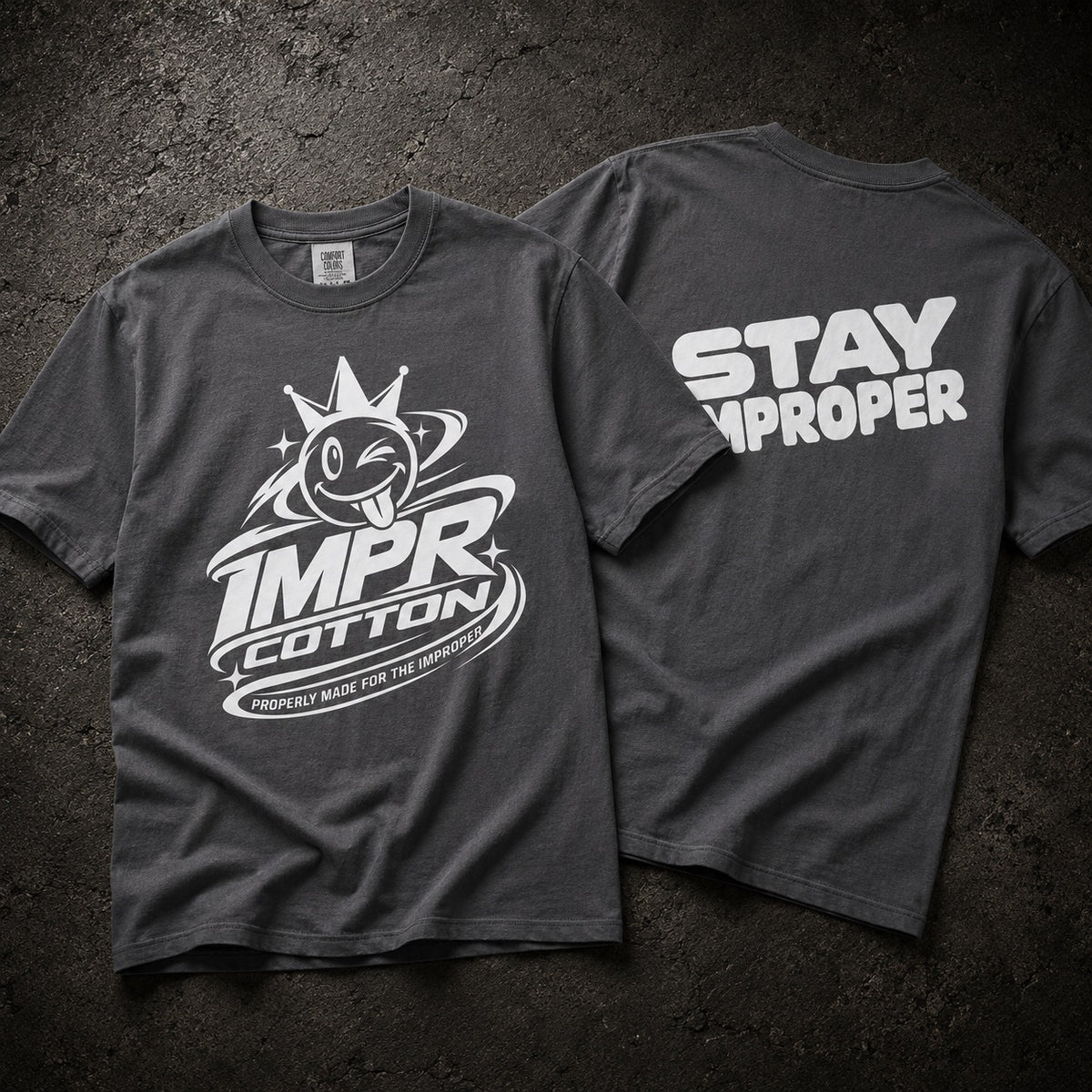 Two dark gray graphic T-shirts laid flat on a textured dark background, showing the front and back. The front has a white cartoon tongue-out logo with the words IMPR Cotton, and the back has large white text reading STAY IMPROPER.