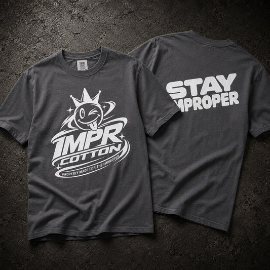 Two dark gray graphic T-shirts laid flat on a textured dark background, showing the front and back. The front has a white cartoon tongue-out logo with the words IMPR Cotton, and the back has large white text reading STAY IMPROPER.