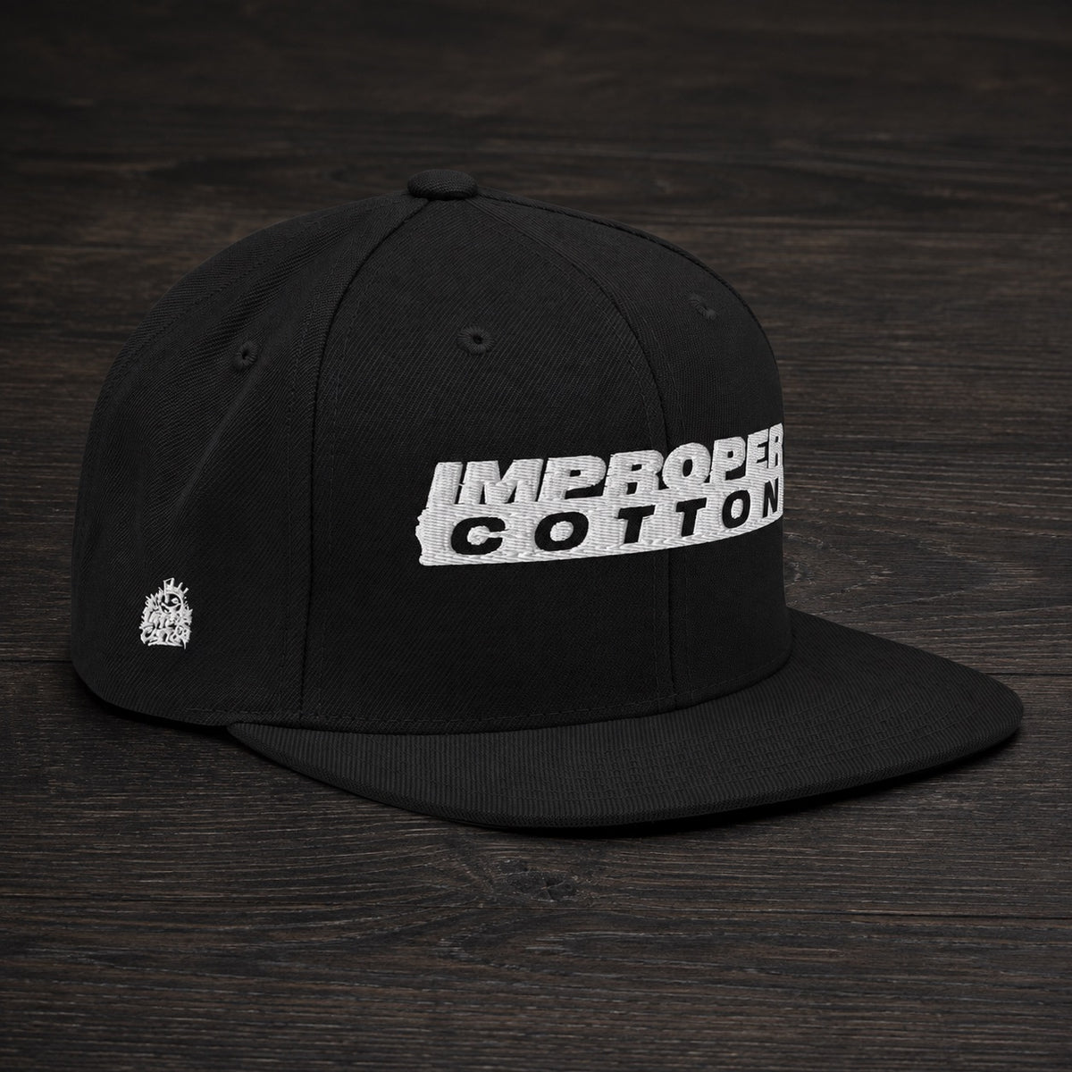 A black flat-brim baseball cap with a white embroidered 'IMPROPER COTTON' logo on the front, displayed on a dark wood background.