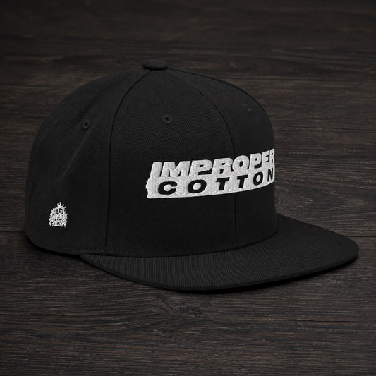 A black flat-brim baseball cap with a white embroidered 'IMPROPER COTTON' logo on the front, displayed on a dark wood background.