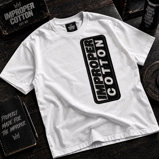 A white short-sleeve T-shirt laid flat on a dark wooden surface, featuring a large black vertical rectangular graphic on the front with the words 'IMPROPER COTTON' in white. The shirt is styled with branded props including a notebook, a sign, and a weight plate.