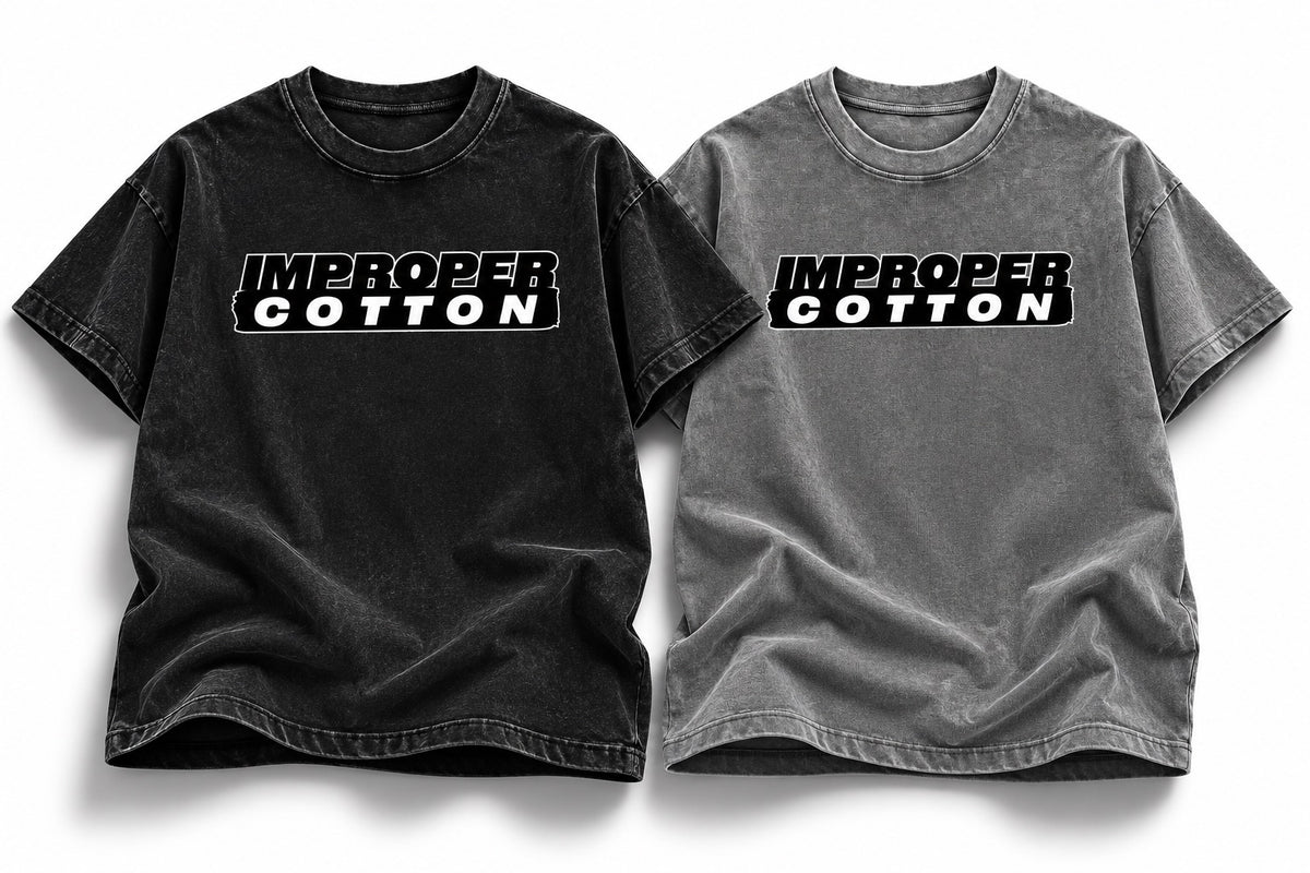Two short-sleeve crewneck t-shirts laid flat side by side on a white background. The left shirt is washed black with a black graphic reading 'IMPROPER COTTON' across the chest, and the right shirt is washed gray with the same black graphic.