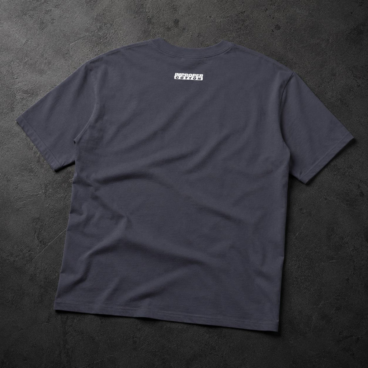 A dark charcoal gray t-shirt laid flat on a textured dark gray surface, featuring a small white rectangular graphic printed on the upper back.