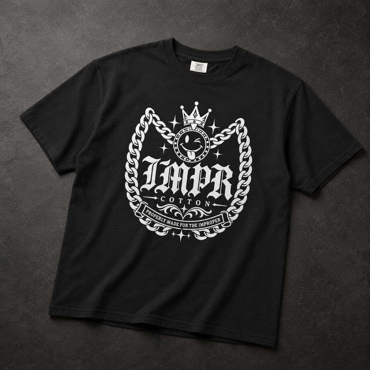 A black short-sleeve t-shirt with a large white graphic print on the front, featuring the text 'IMPR COTTON' inside a chain-link crest with a crown, laid flat on a dark textured background.