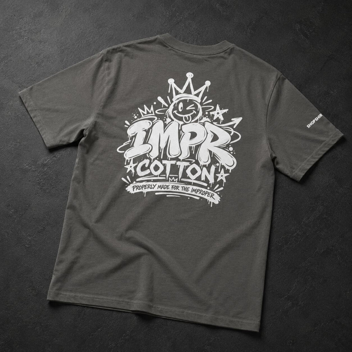 A dark gray t-shirt shown from the back, featuring a large white graphic print with the text 'IMPR COTTON' and a cartoon character wearing a crown. The t-shirt is laid flat on a dark textured background.