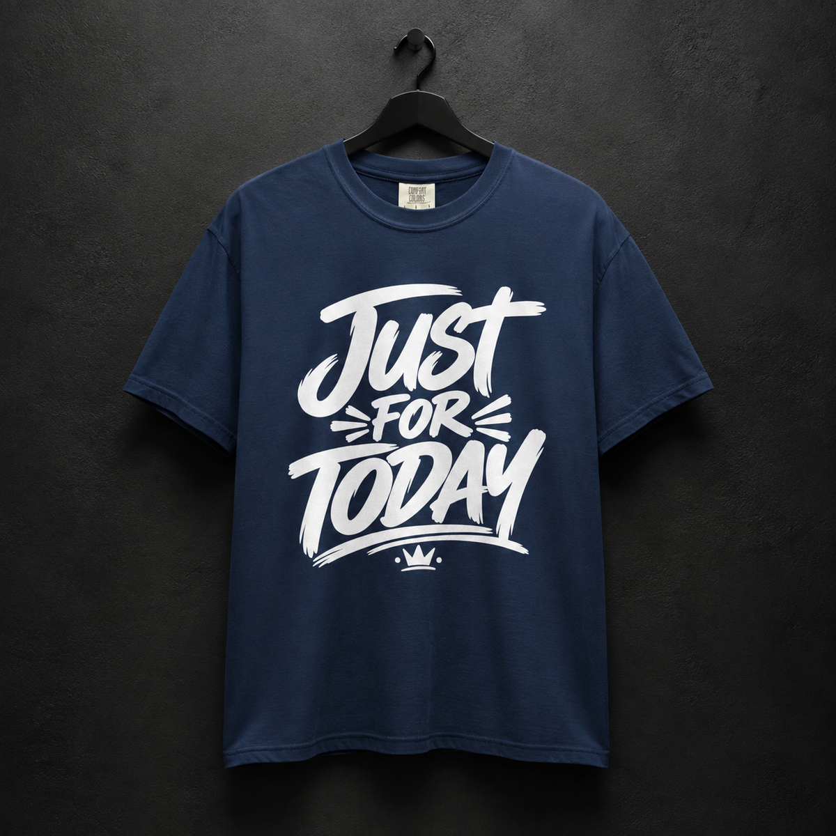 A navy blue short-sleeve t-shirt hanging on a black hanger against a dark textured wall. The shirt features a large white graphic print on the front that reads 'Just For Today' with a small crown logo underneath.