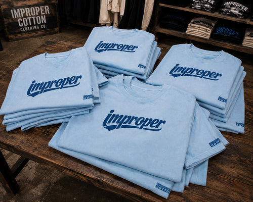 Stacked light blue t-shirts with a dark blue 'improper' logo printed across the chest, displayed on a rustic wooden table inside a retail store.