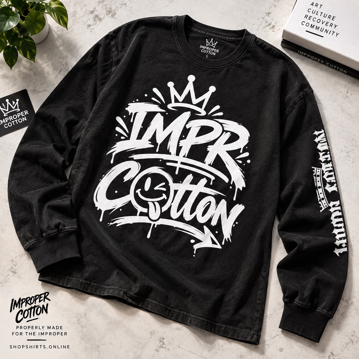 A black long-sleeve graphic t-shirt with a large white 'IMPR Cotton' spray-paint style print on the front, featuring a crown and smiley face detail, laid flat on a light textured surface with decorative props.