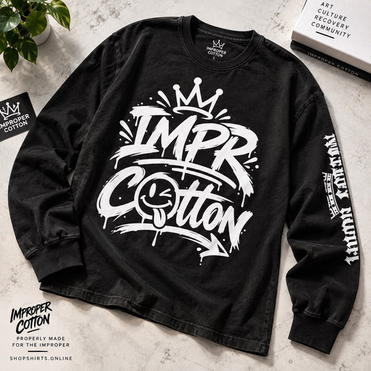A black long-sleeve graphic t-shirt with a large white 'IMPR Cotton' spray-paint style print on the front, featuring a crown and smiley face detail, laid flat on a light textured surface with decorative props.