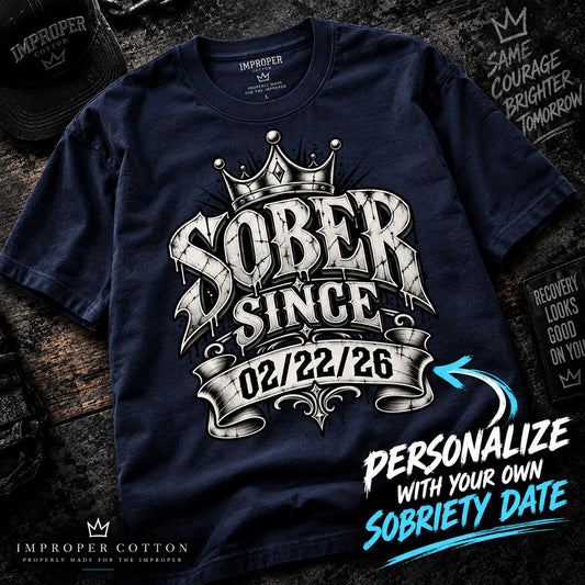 A navy blue t-shirt with a custom graphic reading 'SOBER SINCE 02/22/26' under a crown, styled on a dark wood background with promotional text overlays.