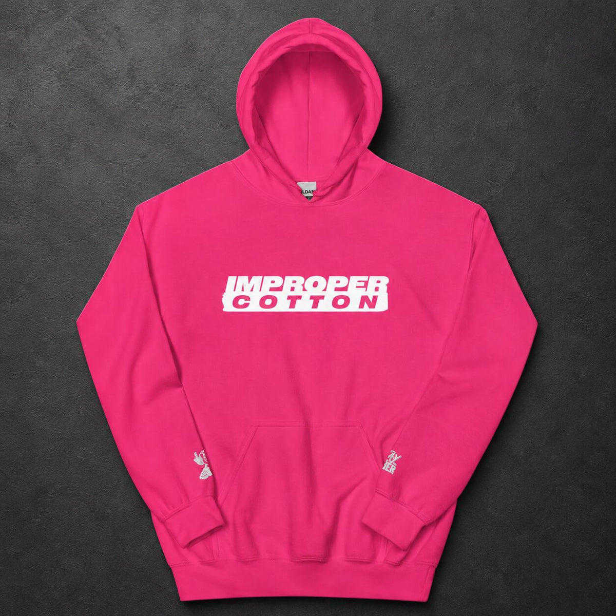 A bright pink pullover hoodie laid flat on a dark textured background. The chest features a white rectangular graphic with the words 'IMPROPER COTTON' in white.