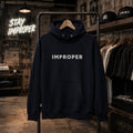A navy blue pullover hoodie with 'IMPROPER' printed in white across the chest, hanging on a black clothing rack inside a styled retail store environment with brick walls and other apparel visible.