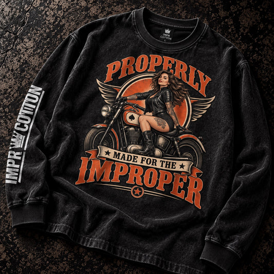 A black long-sleeve t-shirt with a vintage-style biker graphic, laid flat on a dark textured background. The graphic features a woman on a motorcycle with the text 'PROPERLY MADE FOR THE IMPROPER' in orange.