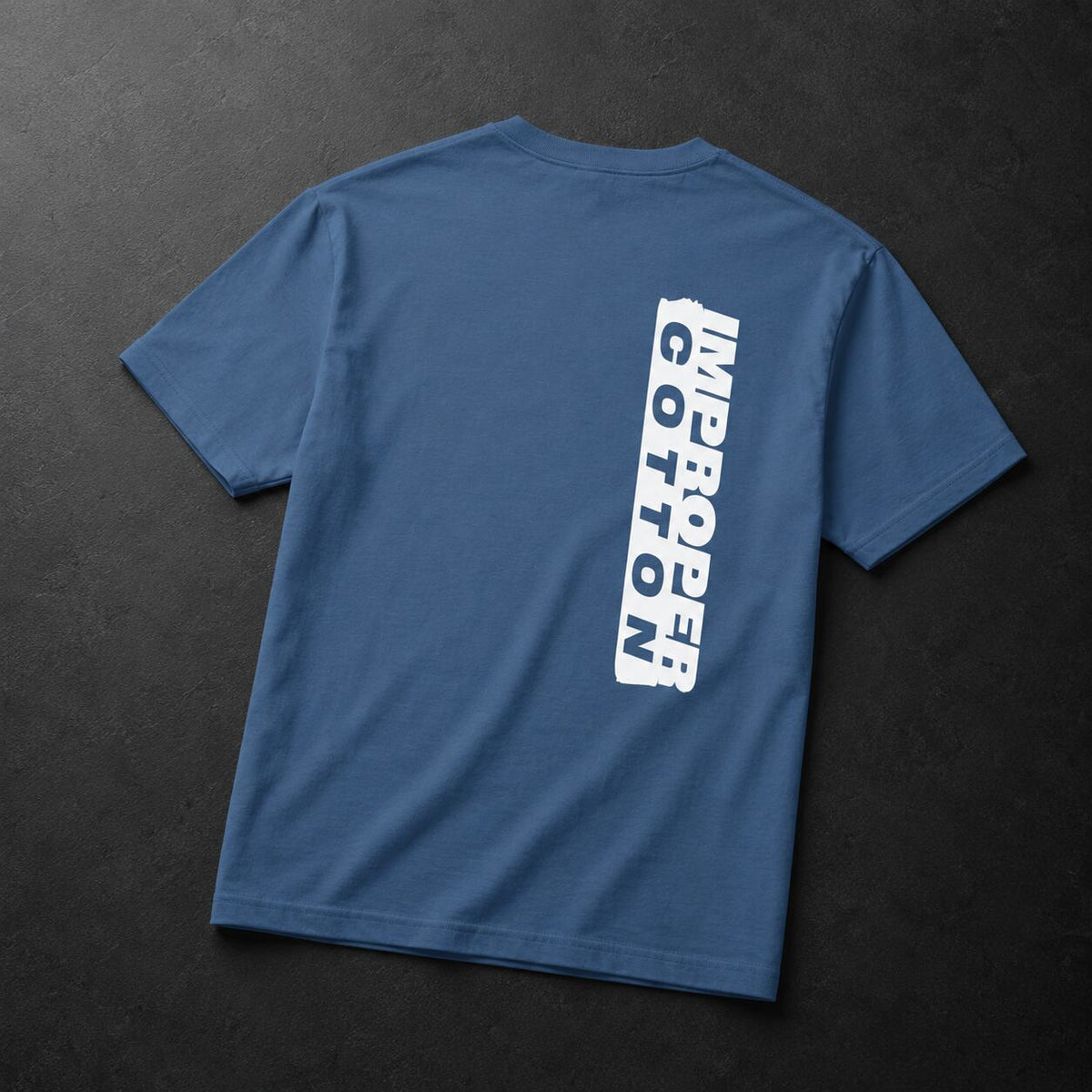 A blue short-sleeve t-shirt shown from the back, laid flat on a dark textured background. The back features a vertical white graphic print reading 'MICROPER COTTON'.
