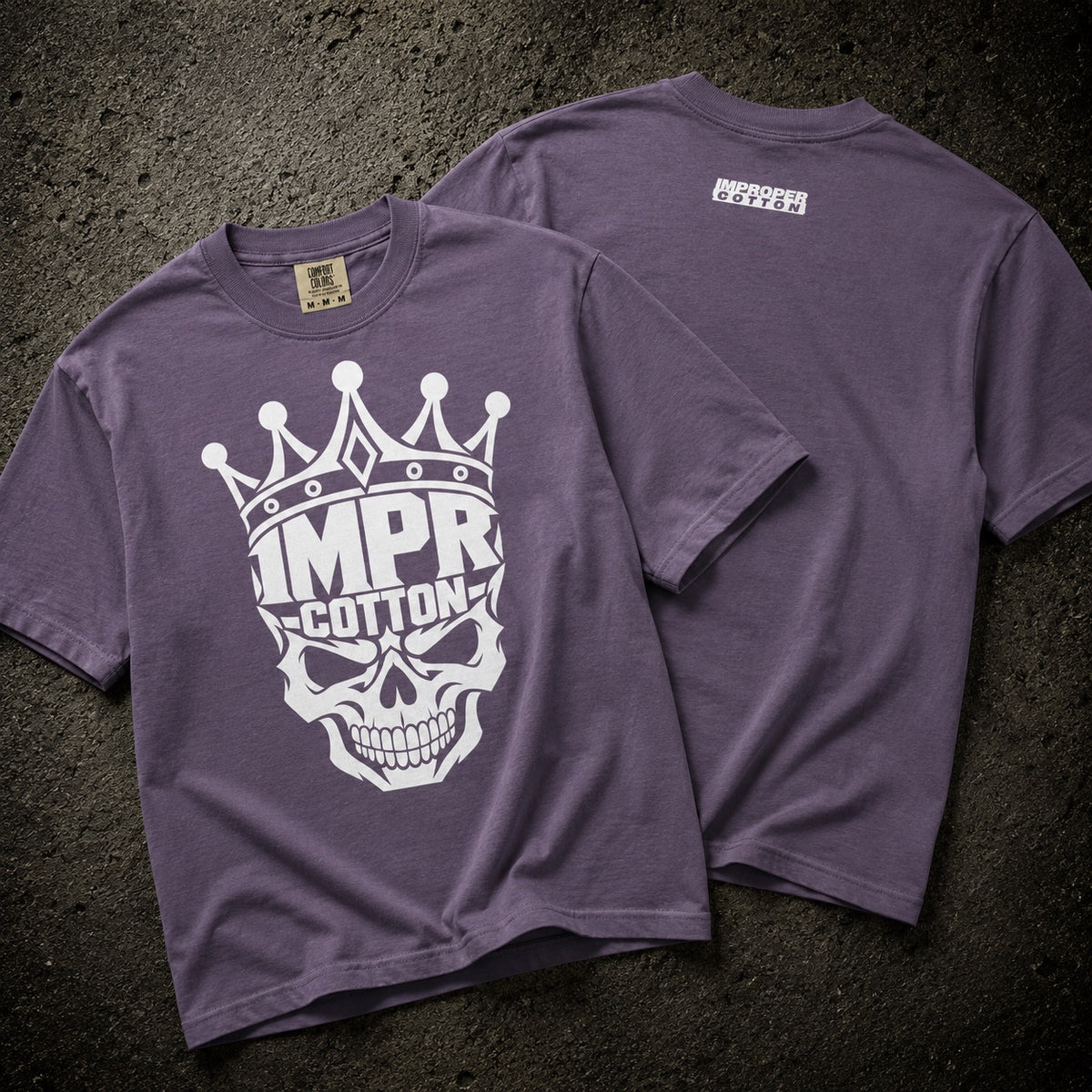 A purple short-sleeve t-shirt shown from both front and back on a dark textured surface. The front features a large white graphic of a skull wearing a crown with the text 'IMPR-COTTON'.