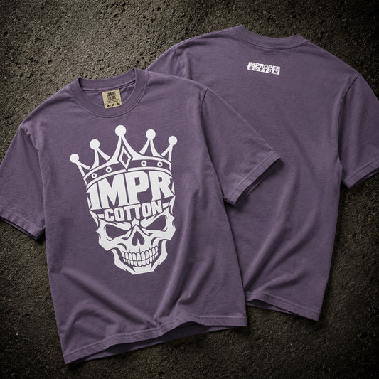 A purple short-sleeve t-shirt shown from both front and back on a dark textured surface. The front features a large white graphic of a skull wearing a crown with the text 'IMPR-COTTON'.