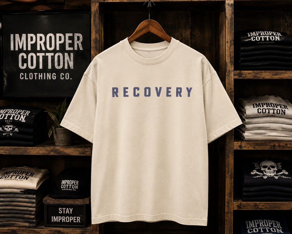 An oversized cream-colored t-shirt with the word 'RECOVERY' printed in navy blue across the chest, hanging on a wooden hanger in front of a rustic dark wood shelving unit filled with other t-shirts.