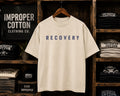An oversized cream-colored t-shirt with the word 'RECOVERY' printed in navy blue across the chest, hanging on a wooden hanger in front of a rustic dark wood shelving unit filled with other t-shirts.