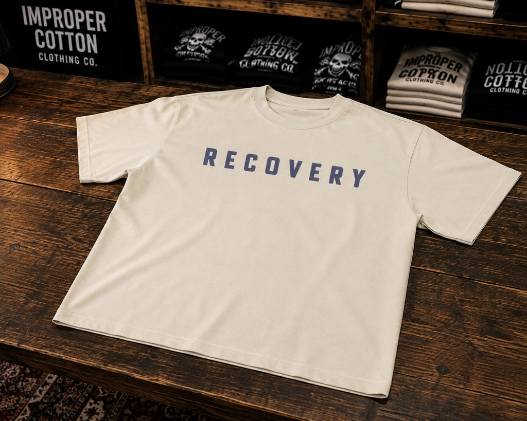 An off-white short-sleeve t-shirt with the word 'RECOVERY' printed in navy blue block letters across the chest, displayed on a dark wooden table with branded merchandise in the background.