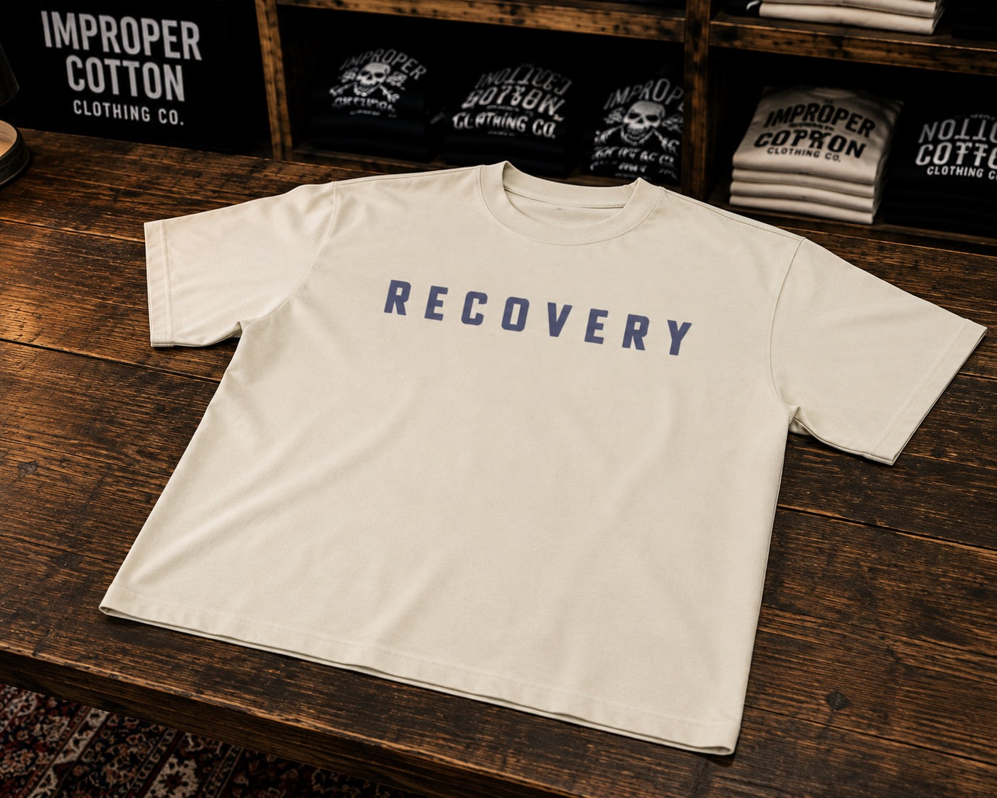 An off-white short-sleeve t-shirt with the word 'RECOVERY' printed in navy blue block letters across the chest, displayed on a dark wooden table with branded merchandise in the background.
