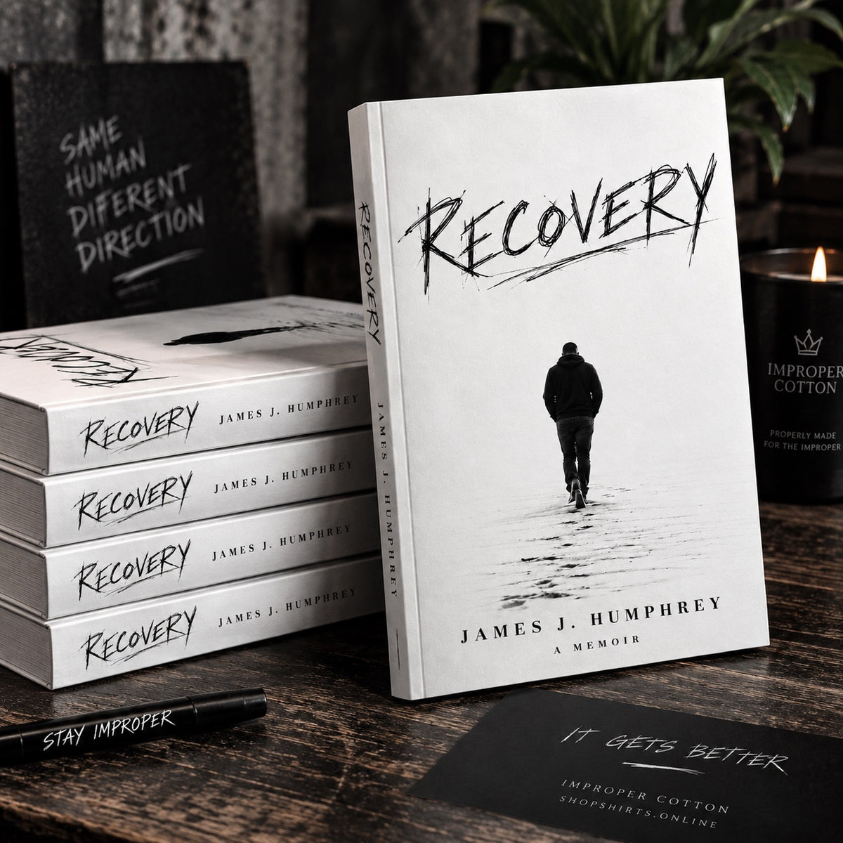 Stack of Recovery A Memoir by James J. Humphrey books on a dark wooden table with one copy standing upright, featuring the minimalist black-and-white cover and Improper Cotton branding a.