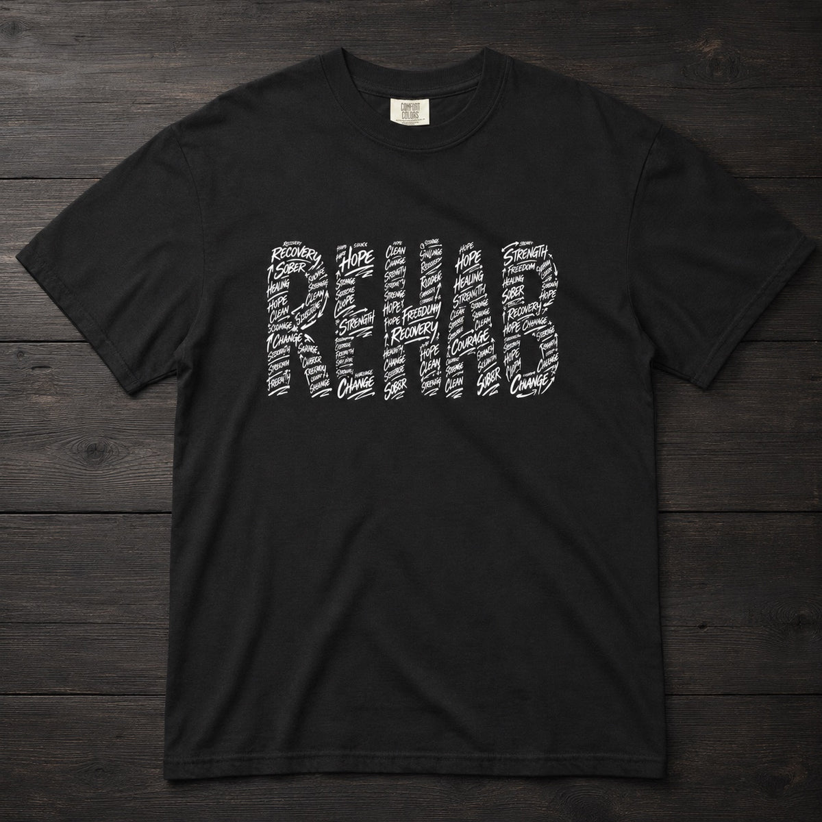 A black short-sleeve t-shirt with the word 'REHAB' printed across the chest in a white typographic pattern, laid flat on a dark wood plank background.