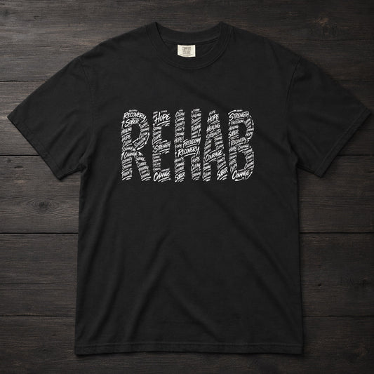 A black short-sleeve t-shirt with the word 'REHAB' printed across the chest in a white typographic pattern, laid flat on a dark wood plank background.