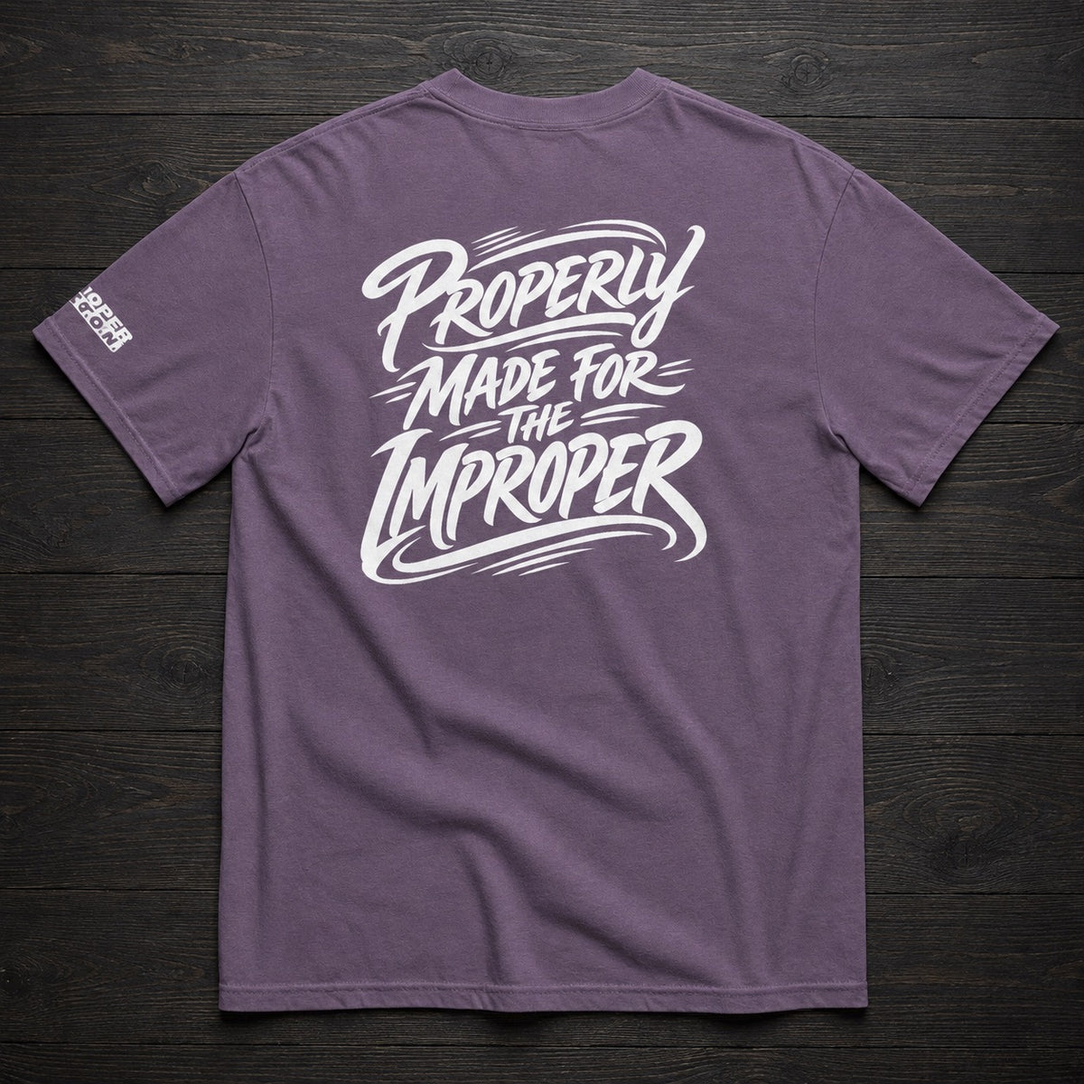 A heathered purple t-shirt laid flat on a dark wood background, featuring a large white graphic on the back that reads 'PROPERLY MADE FOR THE IMPROPER' in a stylized brush font, with a smaller white logo on the left sleeve.