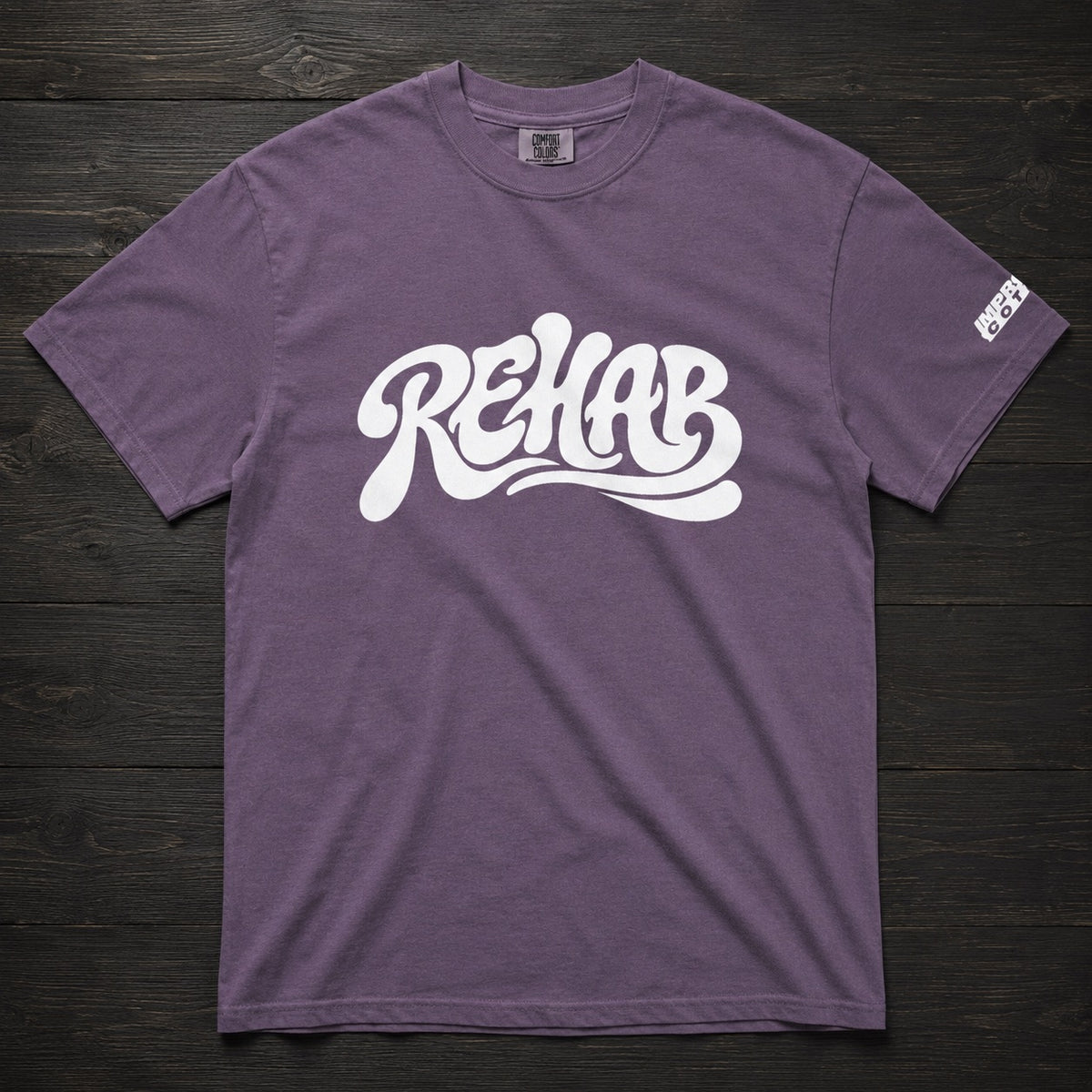 A heathered purple t-shirt with a large white 'REHAB' graphic printed across the chest, laid flat on a dark wood background. A small white brand tag is visible on the right sleeve.