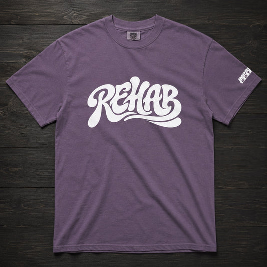 A heathered purple t-shirt with a large white 'REHAB' graphic printed across the chest, laid flat on a dark wood background. A small white brand tag is visible on the right sleeve.