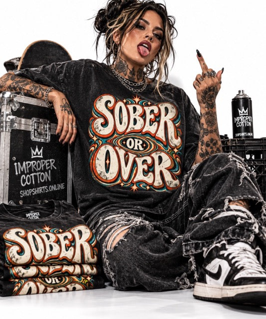 Tattooed female model wearing a black vintage wash SOBER OR OVER retro recovery graphic T-shirt with distressed jeans and streetwear accessories, surrounded by folded shirts and IMPROPER COTTON branding on a clean white background with soft shadow.