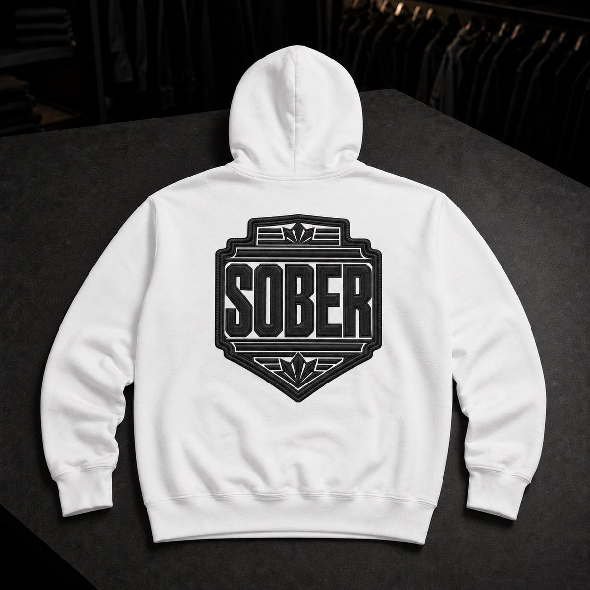 A white pullover hoodie shown from the back, featuring a large black shield-style graphic with the word 'SOBER' in bold letters. The hoodie is laid flat on a dark surface with a blurred clothing rack in the background.