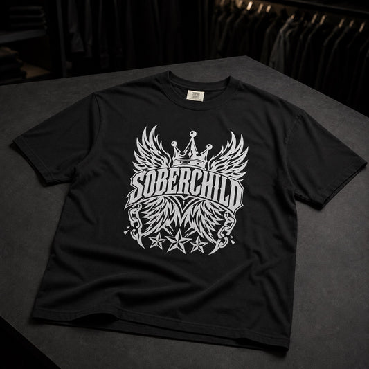 A black short-sleeve t-shirt with a large white graphic on the front featuring wings, a crown, and the text 'SOBERCHILD' and 'MM', displayed on a dark gray surface.