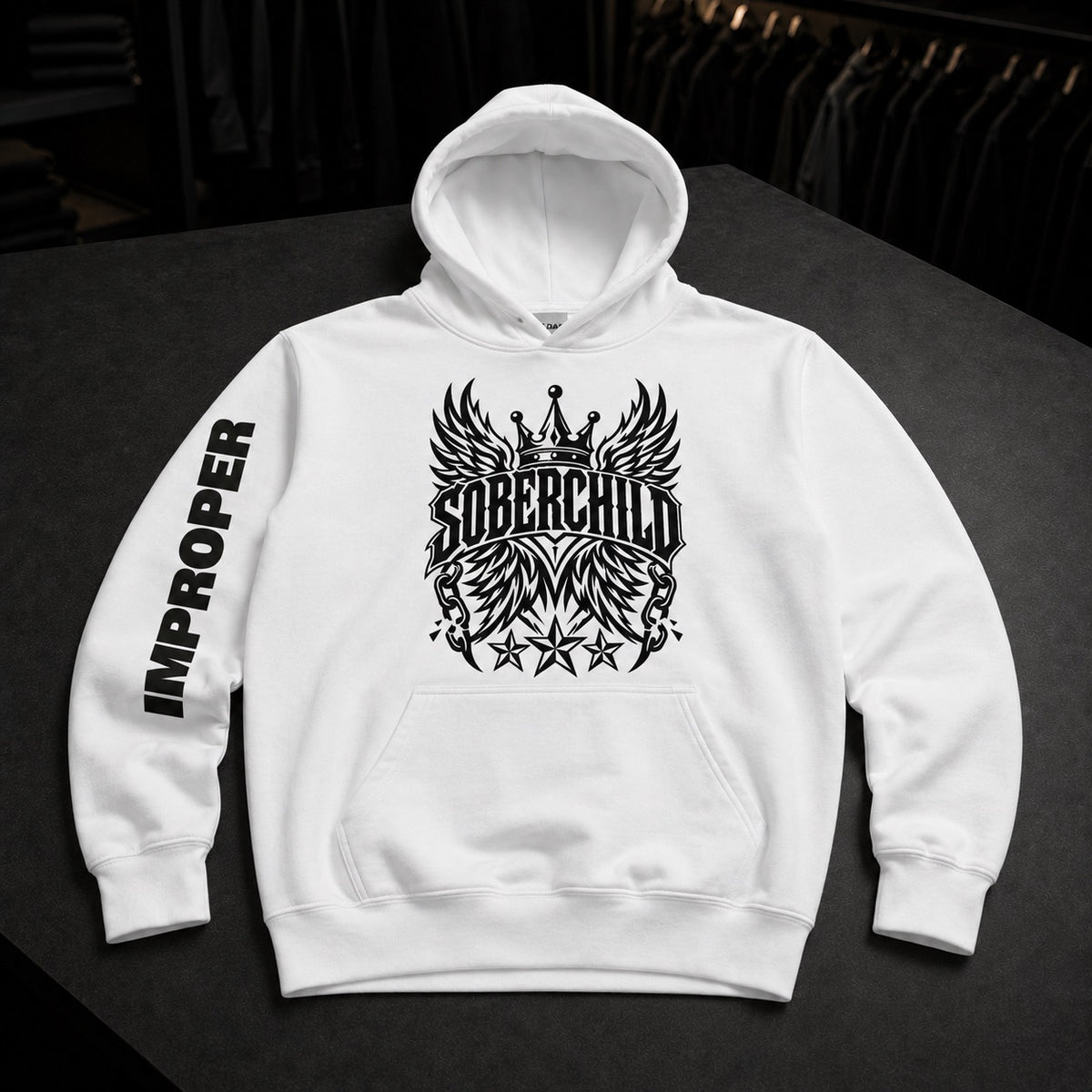 A white pullover hoodie laid flat on a dark surface, featuring a large black winged crown graphic with the word 'SOBERCHILD' across the chest and 'IMPROPER' printed vertically on one sleeve.